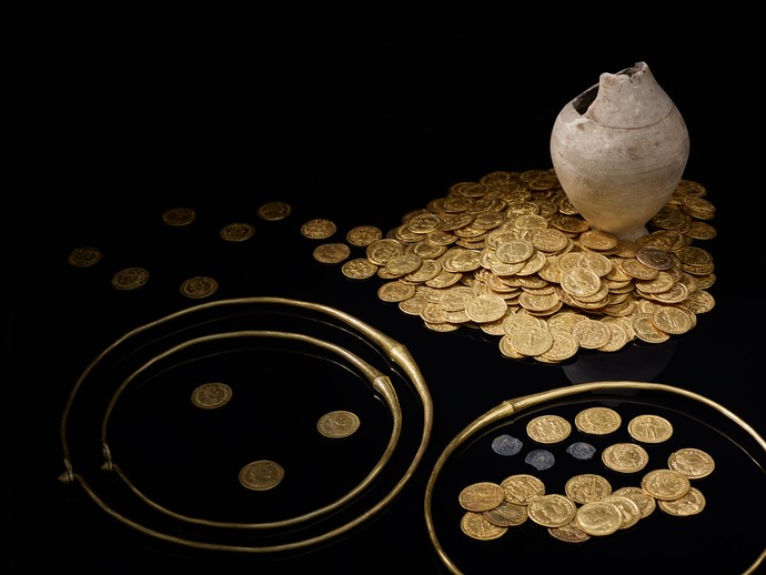 Um einen Tonkrug liegen ein großer Haufen Goldmünzen und drei Goldringe.
