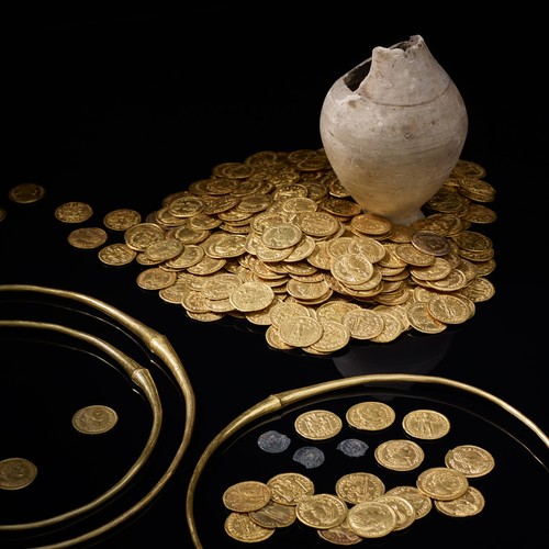Um einen Tonkrug liegen ein großer Haufen Goldmünzen und drei Goldringe.