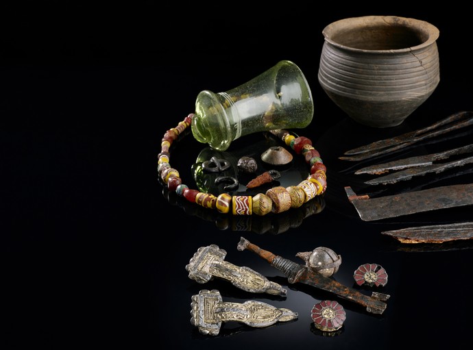 Grabinventar, das mehrere unterschiedliche Gegenstände beinhaltet, darunter ein Glas und Fibeln.